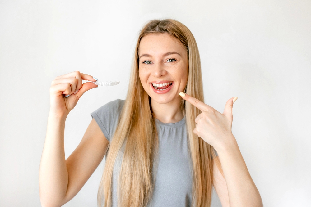 a blonde woman smiling pointing at her teeth while holding invisalign, highlighting Invisalign cost in Rocky point, NY