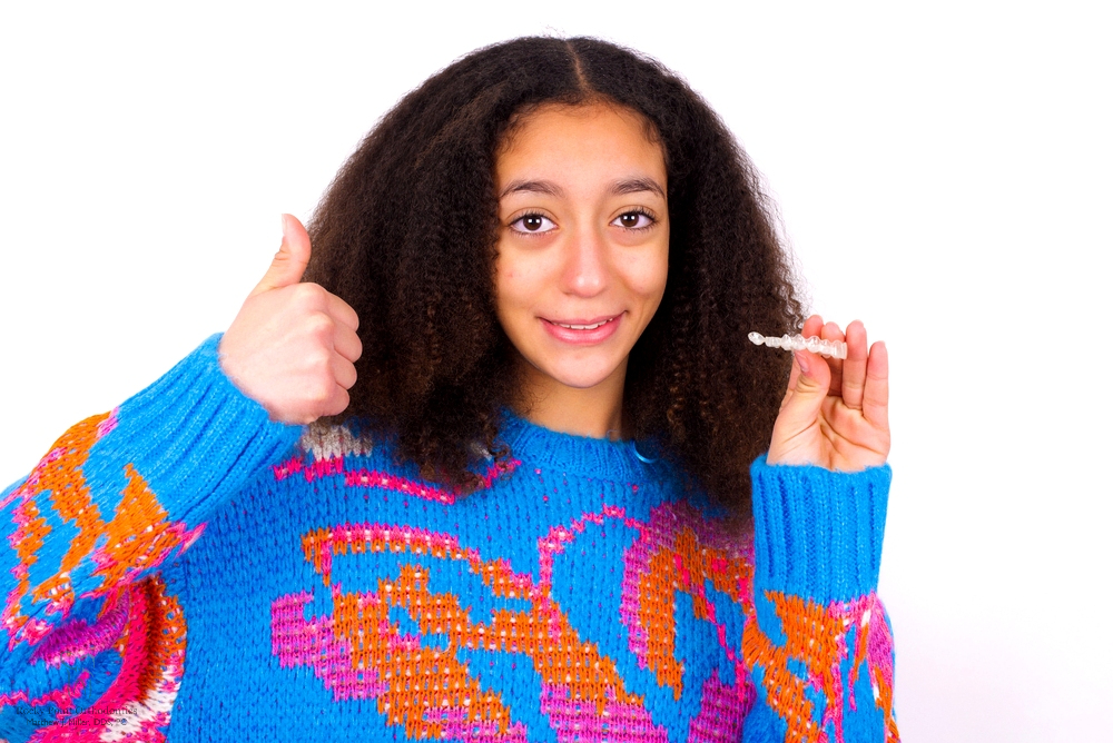 A girl in a colorful sweater holding a clear aligner and giving a thumbs-up, showcasing a modern method for how to fix overbite in Rocky Point, NY.