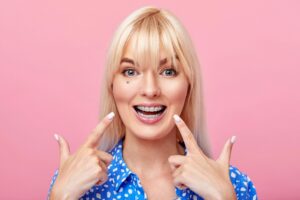 cheerful happy young woman blonde hair gesturing thumb while pointing clear braces - Clear Braces vs. Metal Braces in Rocky Point, NY