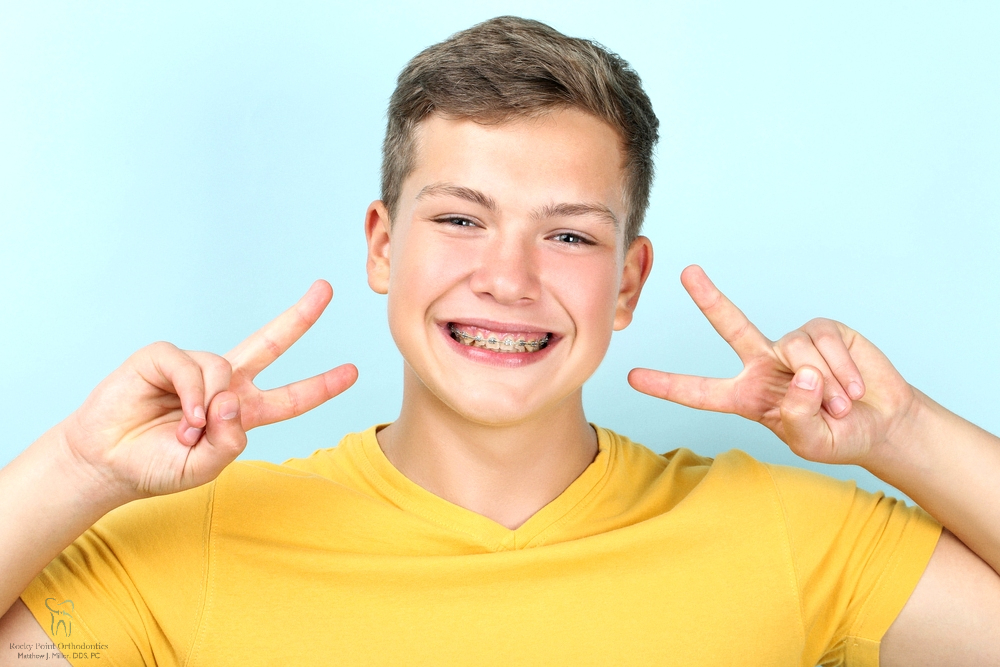 young man metal braces showing two fingers blue background - Invisalign vs Braces Cost in Rocky Point, NY 
